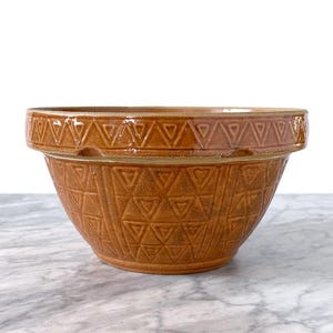 May include: A brown ceramic mixing bowl featuring a wide, flat rim and a textured exterior pattern of repeating triangles. The bowl has a glossy finish and a distinct horizontal band separating the rim from the main body of the vessel.
