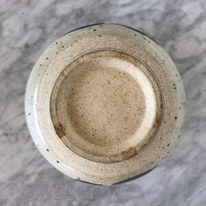 May include: A white ceramic bowl with a speckled glaze and a blue rim. The bowl is shown from the bottom, revealing the unglazed base.