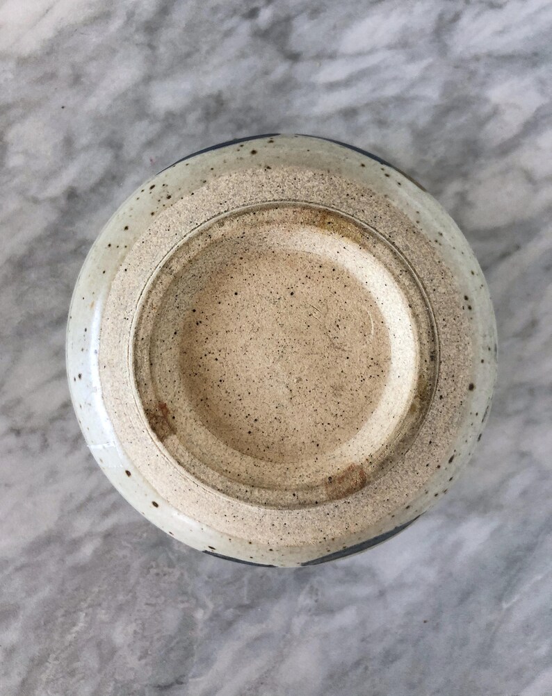 May include: A white ceramic bowl with a speckled glaze and a blue rim. The bowl is shown from the bottom, revealing the unglazed base.