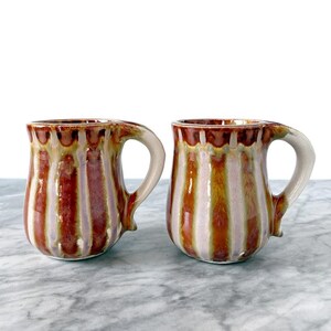 May include: Two ceramic mugs featuring a vertical striped pattern in shades of brown, tan, and light purple. Each mug has a rounded body, a wide rim, and a curved handle extending from the side, finished with a glossy, multi-toned glaze.