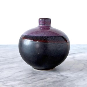 Może przedstawiać: Mały, okrągły ceramiczny wazon z ciemnofioletową górą i czarną podstawą. Wazon ma wąską szyjkę i gładkie, błyszczące wykończenie. Przejście kolorów jest widoczne między ciemnofioletowymi i czarnymi sekcjami.