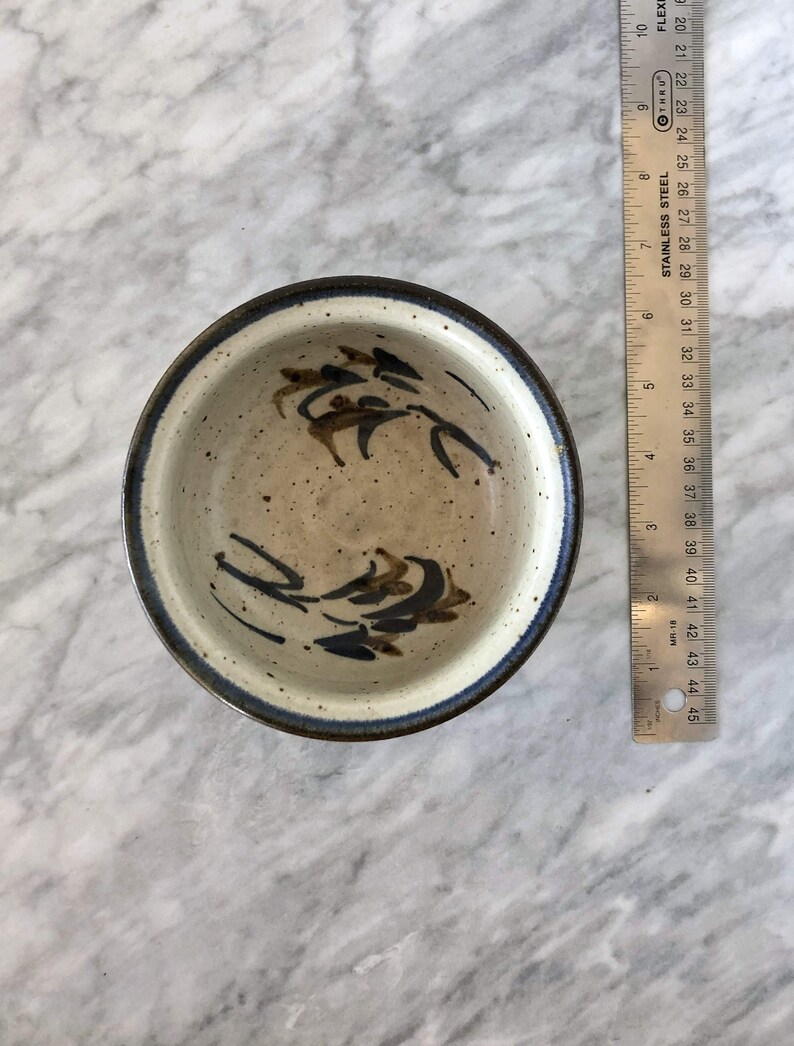 May include: A small, white ceramic bowl with a blue and brown design. The bowl has a speckled glaze and a dark blue rim.