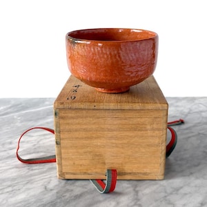 May include: A reddish-orange ceramic tea bowl with a textured surface sits atop a wooden box. The box has a red, gray, and green ribbon attached. The bowl is a traditional Japanese tea ceremony item.