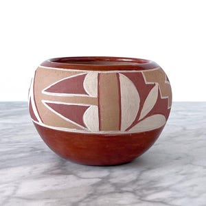 May include: A red clay pot with a geometric design in white and brown. The pot is round and has a wide opening.