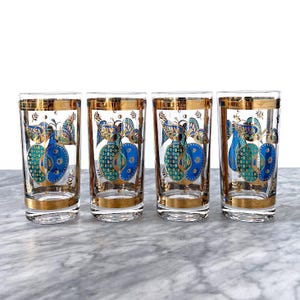 May include: Four clear drinking glasses with gold rims and a gold, blue, and teal apple design. The glasses have a tall, cylindrical shape and are arranged in a row. The apple design is repeated on each glass.