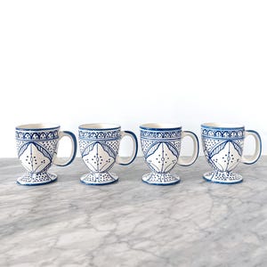 May include: Four white ceramic mugs with blue decorative patterns. Each mug has a handle and a pedestal base. The blue design includes geometric shapes and floral motifs. The mugs are displayed on a marble surface.