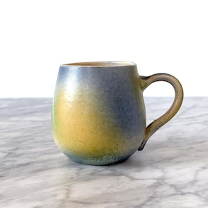 May include: A ceramic mug with a gradient glaze in shades of blue, yellow, and green. The mug has a rounded body and a handle.