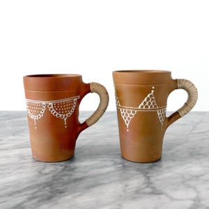 Peut inclure: Deux mugs en terre cuite avec des motifs blancs peints. Les tasses ont des anses tressées. Les mugs cylindriques ont un dessus légèrement évasé. Les motifs blancs sont géométriques et en forme de dentelle. Fond blanc.