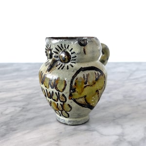 May include: A ceramic owl-shaped pitcher with a small handle. The pitcher is painted in shades of green, brown, and cream, with black detailing. The owl's eyes are metallic, and the design is rustic and handmade.