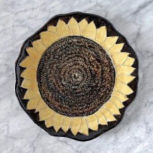 May include: A ceramic plate with a sunflower design. The plate features a dark brown rim and a yellow and brown sunflower pattern in the center. The sunflower petals are yellow, and the center is a textured brown.