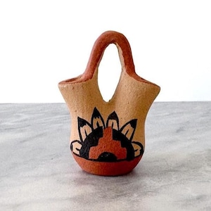 May include: A small, tan-colored ceramic basket with a handle. The basket features a black and red floral design on the front. The handle and base are a darker shade of brown. The basket is likely handmade and has a rustic aesthetic.