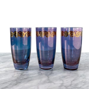 May include: Three tall, elegant drinking glasses. Each glass is a gradient of blue and purple, with a gold, swirling design near the rim. The glasses are displayed on a marble surface.