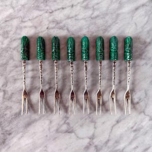 May include: A set of eight silver-toned appetizer forks. Each fork has a two-pronged design and a handle topped with a carved, dark green, cylindrical ornament. The forks are arranged in a row on a light gray, marbled surface.