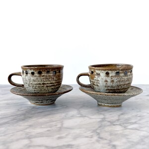 May include: Two ceramic teacups and saucers. The cups and saucers are speckled with brown and white tones. The cups have a handle and a row of dark brown dots near the rim. The saucers are round and have a slightly raised edge.