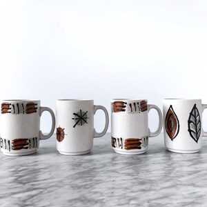 May include: Four white ceramic mugs with brown and gray abstract designs. The mugs have handles and are arranged in a row on a white marble surface.