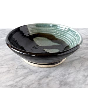 May include: A handmade ceramic bowl with a black exterior and a contrasting interior featuring shades of green and brown. The bowl has a wide, shallow design with a textured rim and a white base. This unique piece is perfect for serving food or as a decorative item.