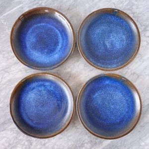 May include: Four round ceramic plates with a deep blue glaze and a brown rim. The plates are arranged in a square pattern, showcasing their glossy finish and unique color variations. These plates are ideal for serving appetizers or desserts.