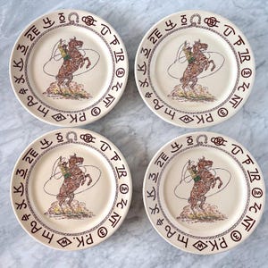 May include: Four cream-colored ceramic plates featuring a central illustration of a cowboy on a rearing horse with a lasso. The rim is decorated with a brown rope pattern and various cattle brand symbols, including the text "The Grazers".
