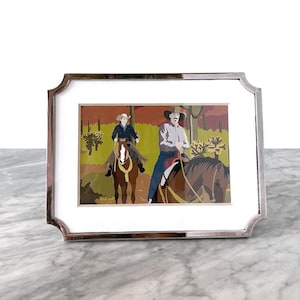 May include: A silver-toned picture frame with a white mat. The framed artwork depicts two people riding horses in a desert landscape. The frame has a decorative, scalloped edge and sits on a marble surface.