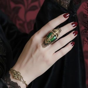 May include: A hand wearing a gold-toned ring with a large green stone and ornate detailing. A matching gold bracelet is also visible. The nails are painted a deep red. The image has a gothic aesthetic.
