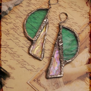 Puede incluir: Un par de pendientes hechos a mano con un diseño único. Cada pendiente presenta un elemento de vidrio verde semicircular y una pieza triangular iridiscente, todo enmarcado en un metal plateado. Los pendientes se exhiben sobre un fondo de papel vintage.