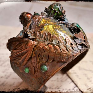 Pode incluir: Uma pulseira de punho ornamentada, de cor bronze com detalhes verdes e dourados. O design incorpora padrões de folhas e pequenas decorações verdes redondas. A pulseira tem uma estética vintage e artística.