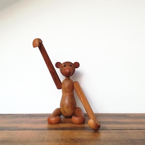 May include: Wooden monkey figurine with articulated limbs. The monkey has a round body, head, and ears, with a long arm raised and another extended. The wood has a warm, natural finish, and the monkey sits on a wooden surface.