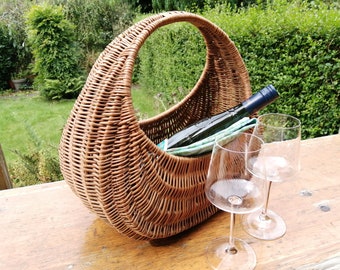 May include: A crescent-shaped woven wicker basket with a high arched handle, containing a dark glass wine bottle and a folded light green patterned cloth, accompanied by two clear stemmed wine glasses on a wooden surface.