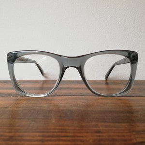 Puede incluir: Gafas grises de ojo de gato con lentes transparentes. Las gafas están sobre una superficie de madera.