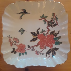 May include: A white ceramic serving dish with a scalloped edge and a floral design. The design features pink flowers, green leaves, and a small bird.