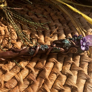 May include: A wooden wand with a purple flower and amethyst crystal at the top. The wand is decorated with green leaves and vines.