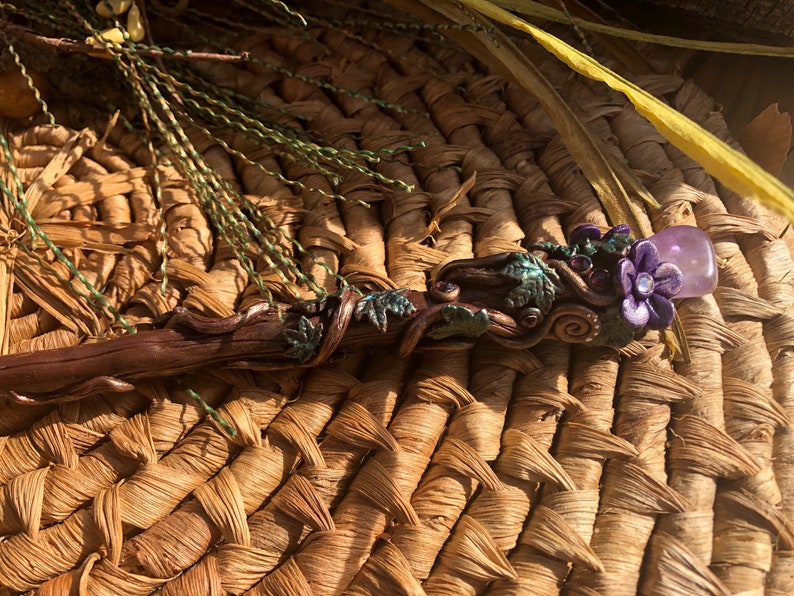 May include: A wooden wand with a purple flower and amethyst crystal at the top. The wand is decorated with green leaves and vines.