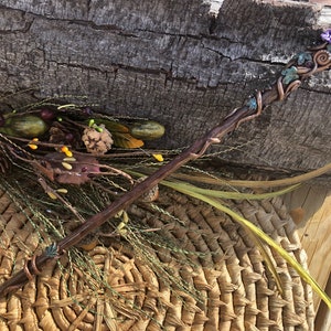 May include: A wooden wand with copper wire wrapped around it, decorated with green and purple polymer clay flowers and a purple crystal at the top.