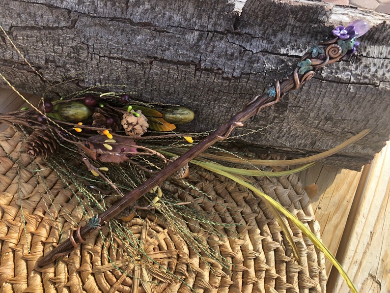 May include: A wooden wand with copper wire wrapped around it, decorated with green and purple polymer clay flowers and a purple crystal at the top.
