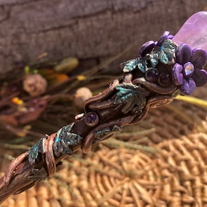 May include: A wooden wand with a purple amethyst stone at the top, decorated with green leaves and purple flowers. The wand is wrapped in copper wire and has small purple gemstones embedded in the wood.