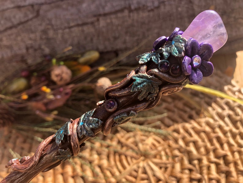 May include: A wooden wand with a purple amethyst stone at the top, decorated with green leaves and purple flowers. The wand is wrapped in copper wire and has small purple gemstones embedded in the wood.