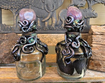 May include: Two glass bottles with black octopus-shaped stoppers and pink crystal tops. The bottles are clear and have a textured surface.
