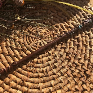 May include: A brown wooden wand with a purple crystal at the top. The wand is decorated with green and brown leaves and vines. The wand is lying on a woven straw mat.