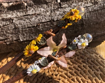 May include: Two wooden wands decorated with polymer clay flowers. One wand has yellow sunflowers and the other has white daisies. Both wands have a clear glass orb at the top.