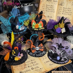 May include: A collection of six miniature black witch hats decorated with colorful tulle, feathers, and themed embellishments like skulls, pumpkins, and character faces. Each hat features hand-painted text on the brim, displayed on a book.