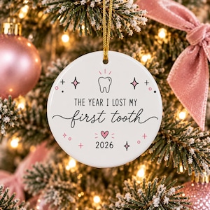 May include: A round white ceramic ornament featuring a black line drawing of a tooth, the text "THE YEAR I LOST MY first tooth", a small pink heart, and the year "2026" in black. The ornament hangs from a gold string against a festive tree.