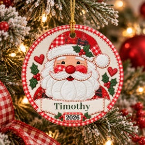 May include: A round Christmas ornament featuring a stylized Santa Claus face with a white beard, red nose, and a red and white checkered hat. The design includes green holly leaves, red berries, small red hearts, and a banner with the name Timothy 2026.