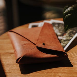 Puede incluir: Una cartera de cuero marrón estilo sobre con cierre de botón dorado. La cartera está doblada y descansa sobre una superficie de madera. La iluminación crea sombras y resalta la textura del cuero. La cartera mide unos 20 cm de largo.