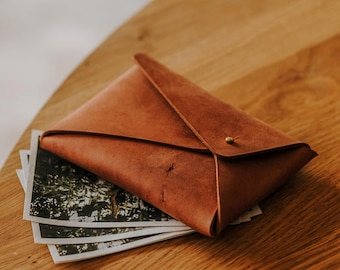 May include: A brown leather envelope sits on a wooden table, with a stack of photos underneath. The photos feature a green and brown landscape.