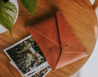 May include: A brown leather envelope with a flap closure and a brass button. The envelope is lying on a wooden table with two photographs underneath it.