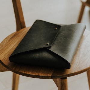 May include: A black leather laptop sleeve with a flap closure and brass accents. The sleeve is sitting on a wooden chair.