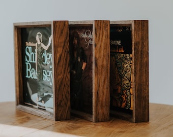 Peut inclure: Trois disques vinyles encadrés avec des pochettes d'album différentes. Les cadres sont en bois foncé avec une finition rustique. Les pochettes d'album présentent une femme en robe bleue, un homme aux cheveux longs et un design abstrait coloré.