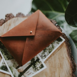 May include: A brown leather envelope with a small gold rivet on the flap, resting on a wooden slice with a blurry photograph of trees underneath.