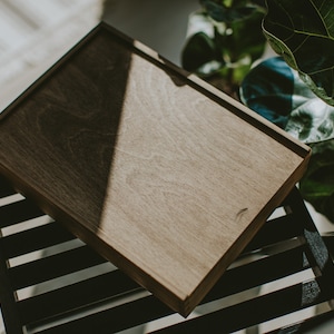 May include: A dark brown wooden box with a smooth surface sits on a black slatted chair. The box is slightly angled, and the light casts a shadow on the surface.