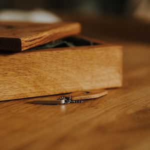 May include: A wooden box with a hinged lid, open to reveal a green interior. A small wooden tag and a silver-colored chain are next to the box. The box and tag are on a wooden surface.